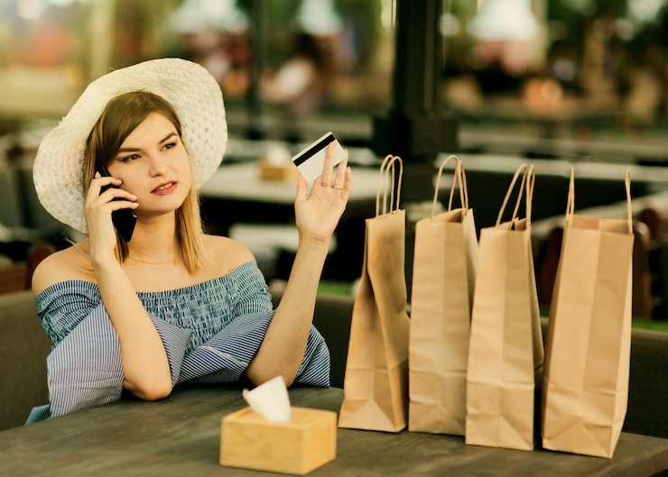 Foto alegre joven adicta a las compras sentado en un café al aire libre mientras habla por teléfono y sostiene una tarjeta bancaria en la mano