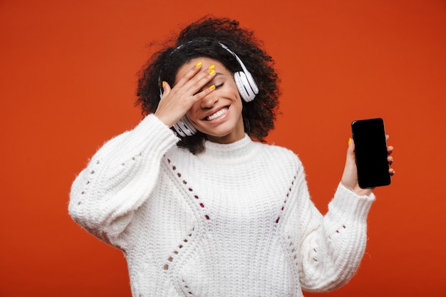 Alegre joven africana atractiva con auriculares inalámbricos que se encuentran aisladas sobre la pared roja, mostrando el teléfono móvil con pantalla en blanco