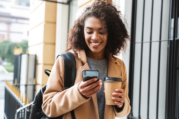 Alegre joven africana vistiendo abrigo caminando al aire libre, sosteniendo la taza de café para llevar, mediante teléfono móvil