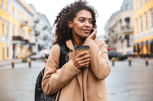 Alegre joven africana vistiendo abrigo caminando al aire libre, sosteniendo la taza de café para llevar