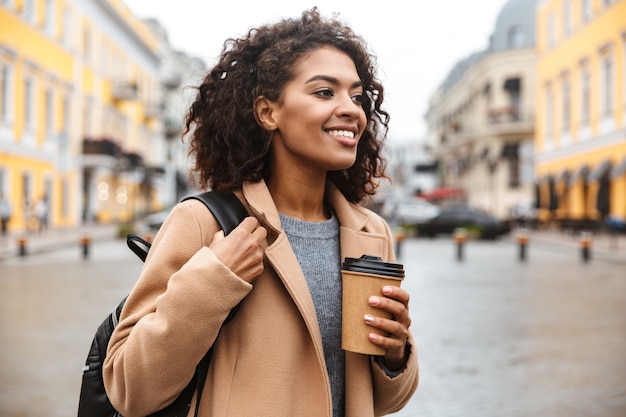 Foto alegre joven africana vistiendo abrigo caminando al aire libre, sosteniendo la taza de café para llevar