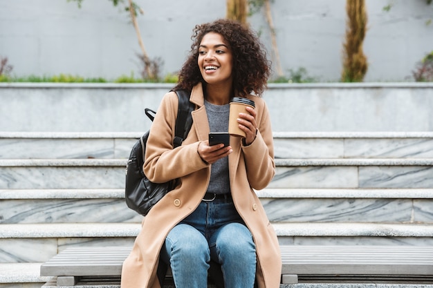 Alegre joven africana vistiendo abrigo sentado al aire libre, sosteniendo la taza de café para llevar