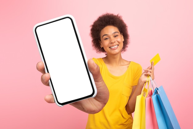 Alegre joven afroamericana rizada con bolsas con compras con tarjeta de crédito muestra el teléfono inteligente con pantalla en blanco