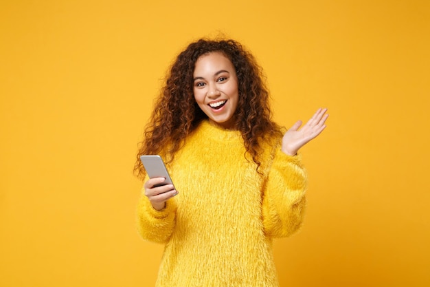 Alegre joven afroamericana con suéter de piel posando aislada en un fondo naranja amarillo, retrato de estudio. Concepto de estilo de vida de las personas. Simulacros de espacio de copia. Sosteniendo el teléfono móvil, extendiendo las manos.