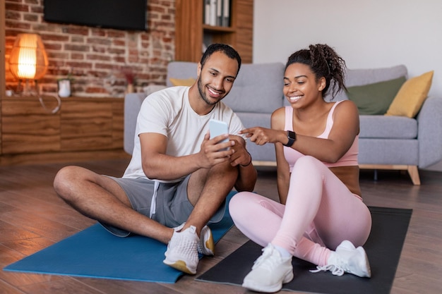 Alegre joven afroamericano marido y mujer en ropa deportiva ver video en el teléfono inteligente en la alfombra