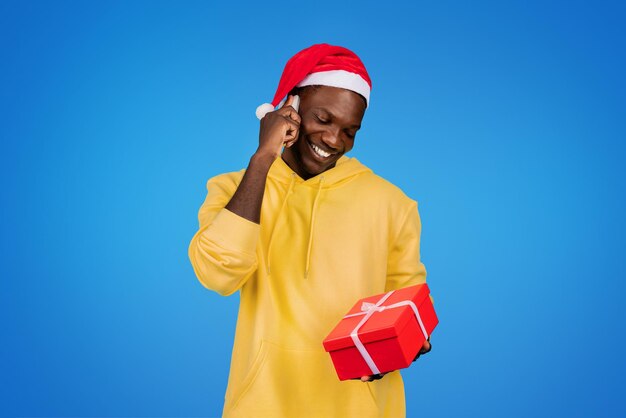 Alegre joven afroamericano con sudadera y gorro de Papá Noel llama por teléfono mostrar caja de regalo