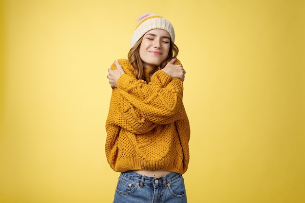 Alegre joven alegre y encantadora que se siente afortunada feliz abrazándose a sí misma como un nuevo suéter cálido y cómodo inclinando la cabeza hacia el hombro con los ojos cerrados sonriendo ampliamente abrazando su propio cuerpo, fondo amarillo