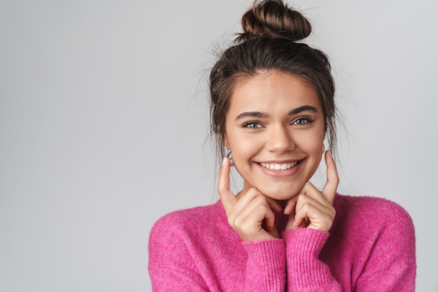 Foto alegre joven apuntando con el dedo a sus mejillas y sonriendo aislado sobre pared gris