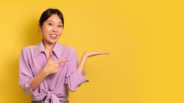 Alegre joven asiática apuntando a un objeto invisible en su palma sobre el fondo amarillo del estudio