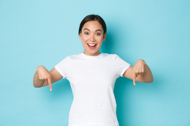 Alegre joven asiática con camiseta blanca apuntando con el dedo hacia abajo y sonriendo emocionada, luciendo optimista mientras muestra la pancarta, ofrece promoción de descuento especial, de pie con fondo azul.