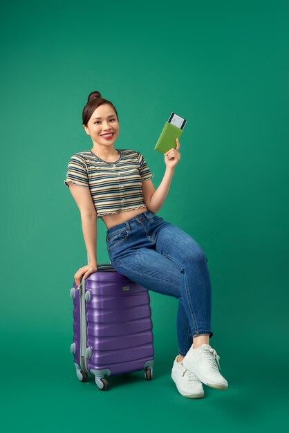 Alegre joven asiática sentada en la maleta y sosteniendo el pasaporte, billete de avión para viajar sobre verde.