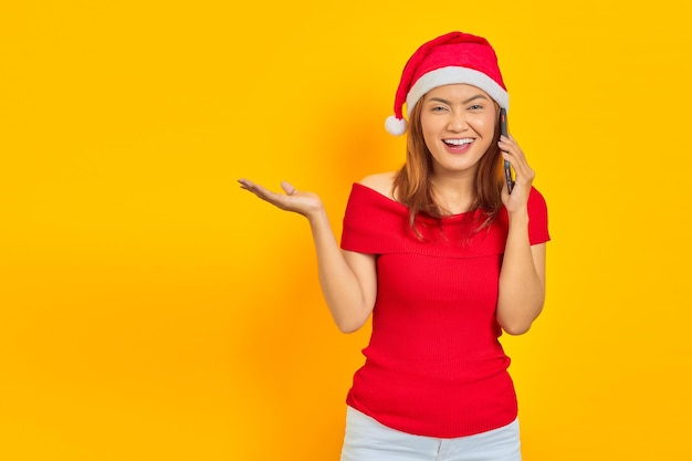 Alegre joven asiática con sombrero de Navidad hablando por teléfono móvil mientras apunta hacia el lado