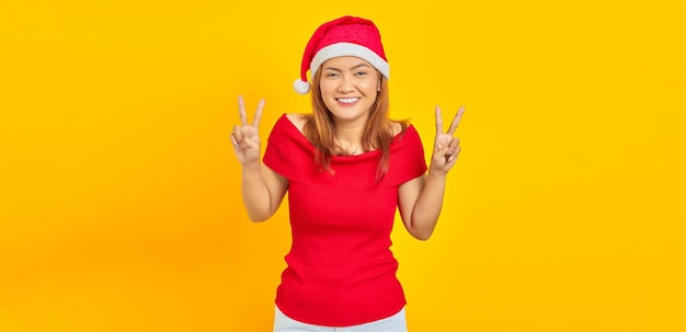 Alegre joven asiática con sombrero de Navidad mostrando el signo de la paz sobre fondo amarillo