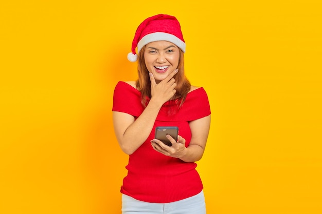 Alegre joven asiática con sombrero de Navidad con smartphone con el dedo en la barbilla