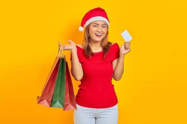 Alegre joven asiática con sombrero de Santa Claus sosteniendo bolsas de la compra y mostrando la tarjeta de crédito