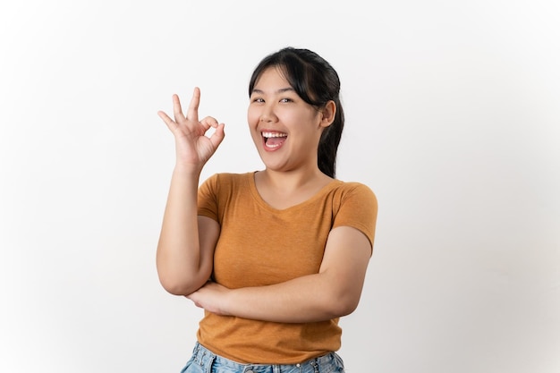 La alegre joven asiática sonriendo y mostrando el signo de la mano OK de pie sobre un fondo blanco