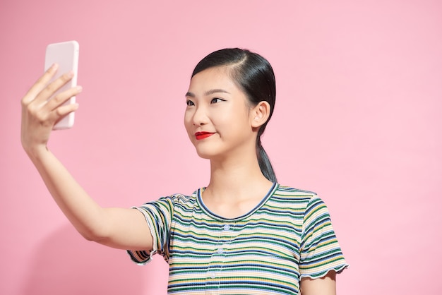 Alegre joven asiática sosteniendo teléfono móvil y haciendo selfie en rosa