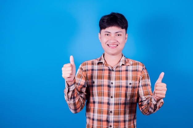 Alegre joven asiático apuesto en camisa a cuadros sobre fondo azul, muestra el pulgar hacia arriba en señal de aprobación