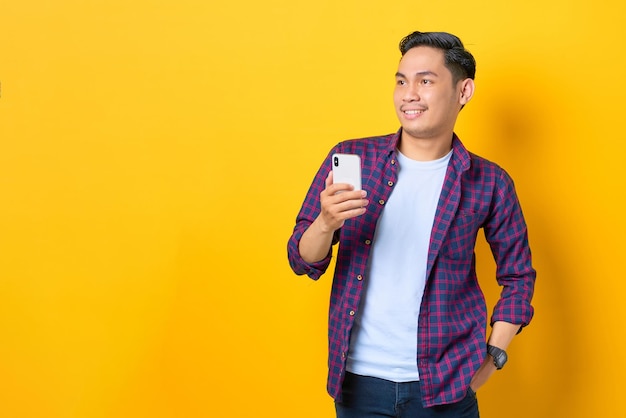 Alegre joven asiático con camisa a cuadros usando un teléfono móvil y mirando hacia otro lado en el espacio de la copia aislado en el fondo amarillo