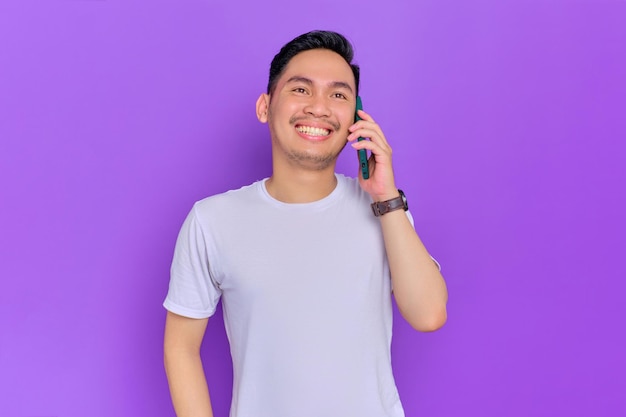 Alegre joven asiático con camiseta blanca hablando por teléfono móvil aislado de fondo morado