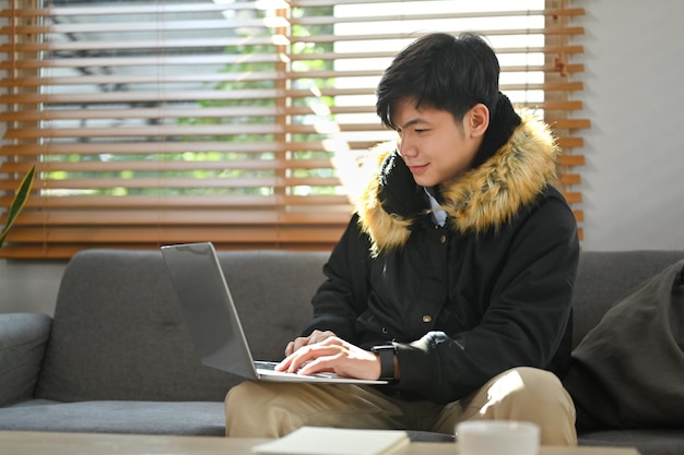 Alegre joven asiático navegando por sitios web de Internet o comprando en línea en una computadora portátil en una acogedora sala de estar