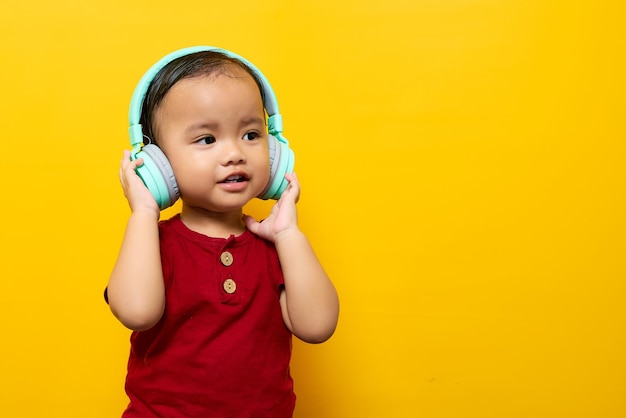 Alegre joven asiático niño de 2 años con camiseta roja Escuchando música favorita con auriculares aislados en fondo amarillo Concepto de estilo de vida familiar del amor del Día de la Madre