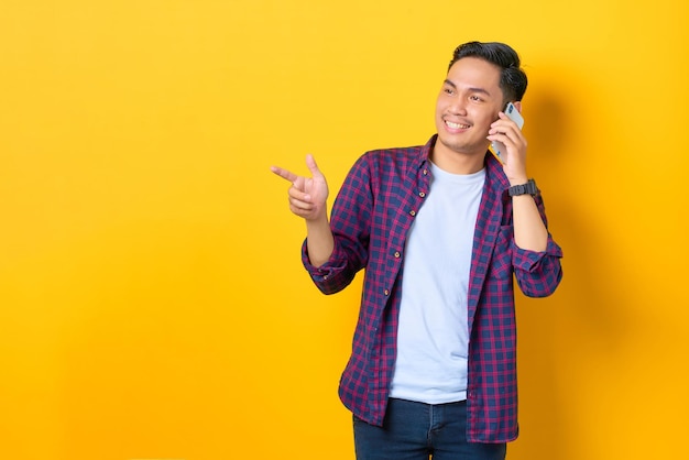 Alegre joven asiático con pantalones a cuadros hablando por teléfono móvil y señalando con el dedo el espacio de copia aislado en el fondo amarillo