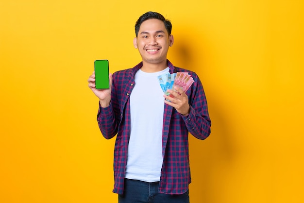 Alegre joven asiático con pantalones a cuadros mostrando un teléfono móvil con pantalla en blanco y sosteniendo billetes de dinero aislados en un fondo amarillo