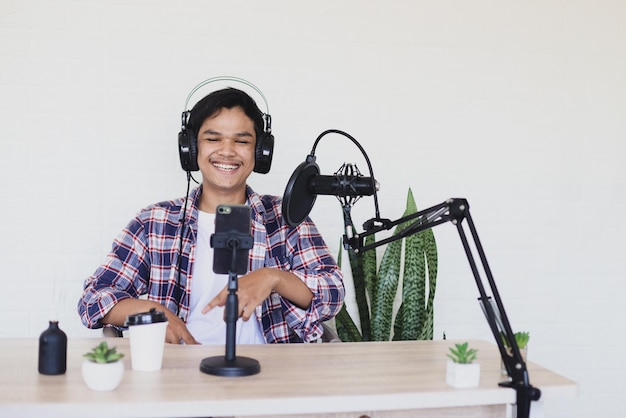 Alegre joven asiático sonriendo durante la filmación de vlog usando un teléfono inteligente y un podcast usando un micrófono en el soporte