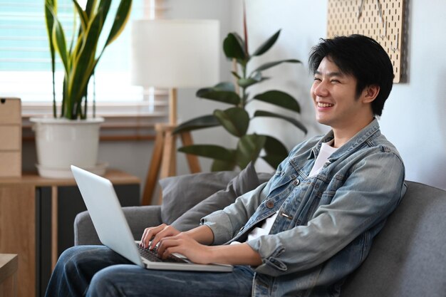 Alegre joven asiático usando computadora portátil mientras está sentado en el sofá en casa