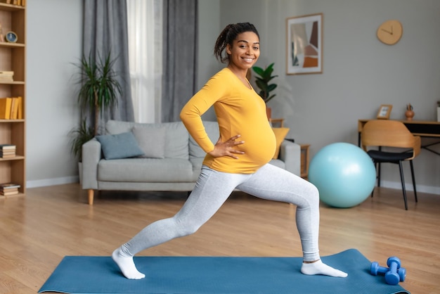 Alegre joven atleta negra embarazada con gran barriga haciendo ejercicios de piernas y practicando yoga