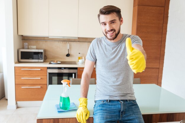 Alegre joven atractivo en guantes de goma amarillos limpiando su piso y mostrando los pulgares para arriba