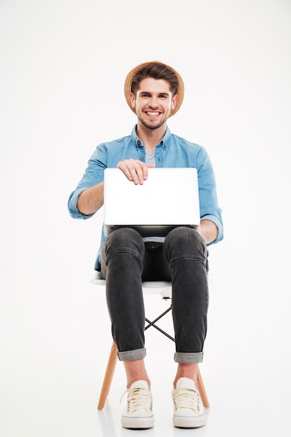 Alegre joven atractivo con sombrero sentado y usando laptop