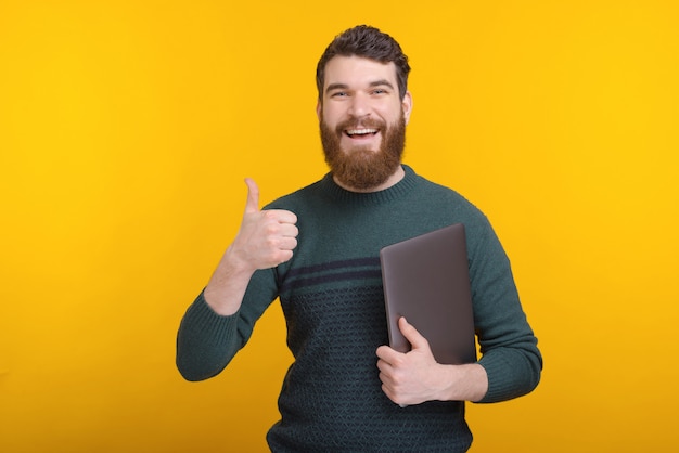 Alegre joven con barba mostrando pulgares arriba gesto y sosteniendo su computadora portátil