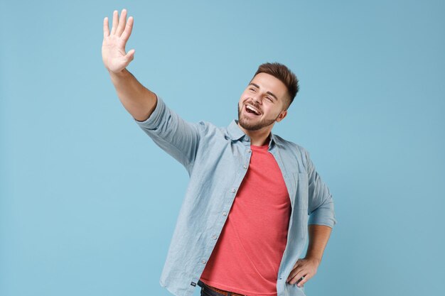 Foto alegre joven barbudo con camisa informal posando aislado en un retrato de estudio de fondo azul pastel. concepto de estilo de vida de las personas. simulacros de espacio de copia. saludando y saludando con la mano cuando alguien se da cuenta.