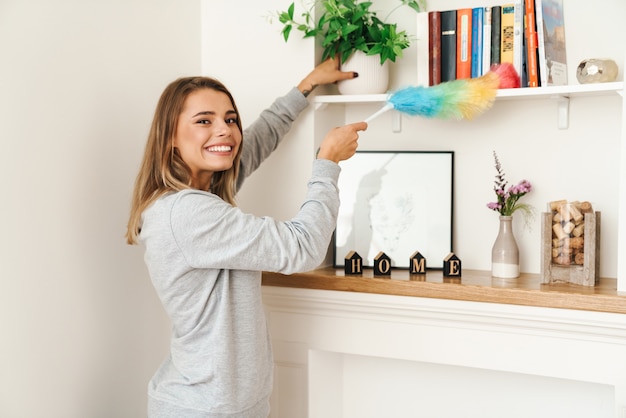 Alegre joven y bella mujer ama de casa sonriendo y haciendo quehaceres domésticos con plumero en la sala de estar