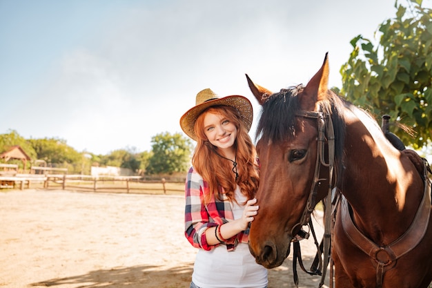 Alegre joven y bella mujer vaquera de pie con su caballo en la aldea