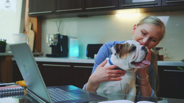 Alegre joven con bulldog cachorro sentado frente a la computadora portátil en la cocina