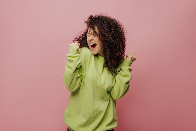 Alegre joven caucásica aprieta los puños en triunfo cierra los ojos de felicidad sobre fondo rosa Morena con rizos tiene una sonrisa con dientes viste ropa informal Concepto de éxito