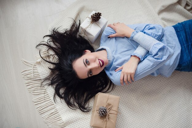 Alegre joven caucásica en jeans se sienta en el suelo cerca del árbol de Navidad en una acogedora habitación decorada.