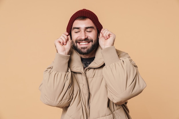 Alegre joven en chaqueta de invierno sonriendo y poniéndose el sombrero aislado sobre pared beige