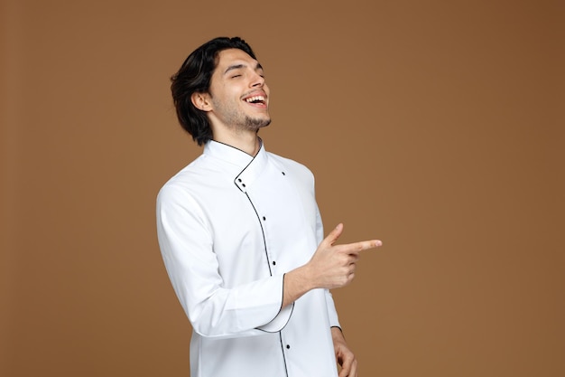 alegre joven chef uniformado apuntando a un lado riéndose con los ojos cerrados aislado de fondo marrón