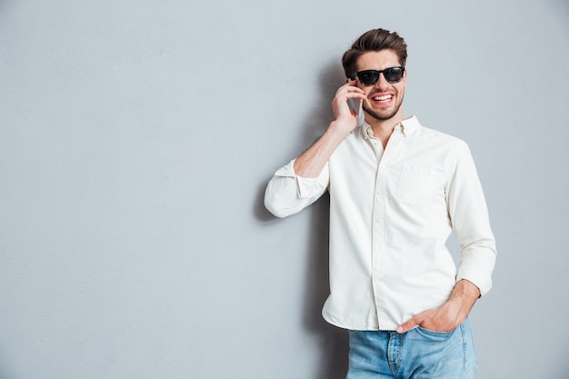 Alegre joven confiado en camisa blanca y gafas de sol hablando por teléfono celular