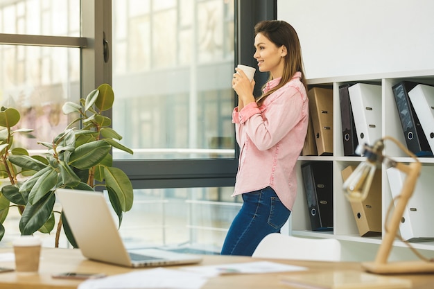 Foto alegre joven empresaria en la oficina tomando café mientras está de pie junto a la ventana en la oficina