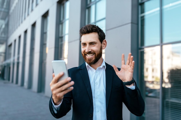 Alegre joven empresario europeo confiado con barba en traje mira la cámara web del teléfono agitando la mano saludando al cliente