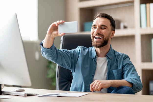 Alegre joven empresario haciendo videollamadas en un teléfono inteligente mientras está sentado en la oficina