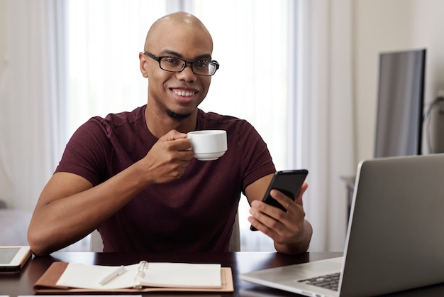 Alegre joven empresario negro bebiendo una taza de café y revisando mensajes y notificaciones en su teléfono inteligente durante un breve descanso en el trabajo