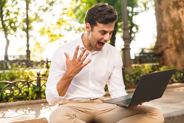 Alegre joven empresario sentado al aire libre, trabajando en equipo portátil, celebrando el éxito