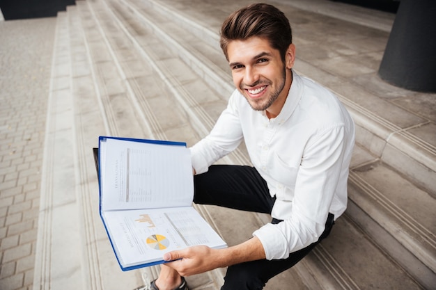 Alegre joven empresario sentado en las escaleras y leyendo el plan de negocios en la carpeta