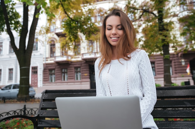 Alegre joven encantadora sonriendo y usando la computadora portátil en el parque